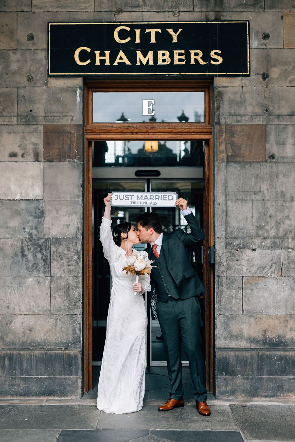 A wedding couple poses for romantic photos in front of a historic stone building with vintage 'City Chambers' signage.