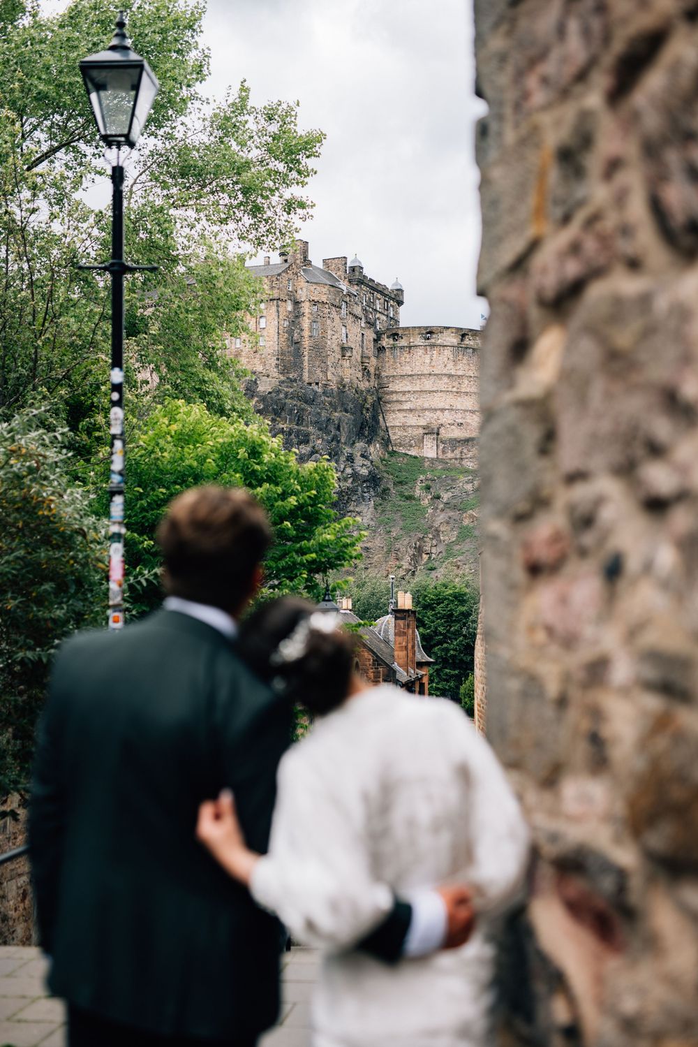 A couple gazes at Edinburgh Castle while standing on a historic cobblestone street with a vintage lamppost.