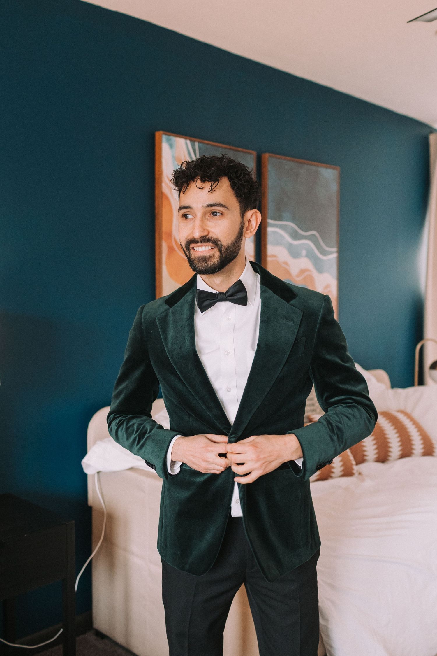 A groom adjusts his black tuxedo jacket while getting ready in an elegant hotel room with teal walls.