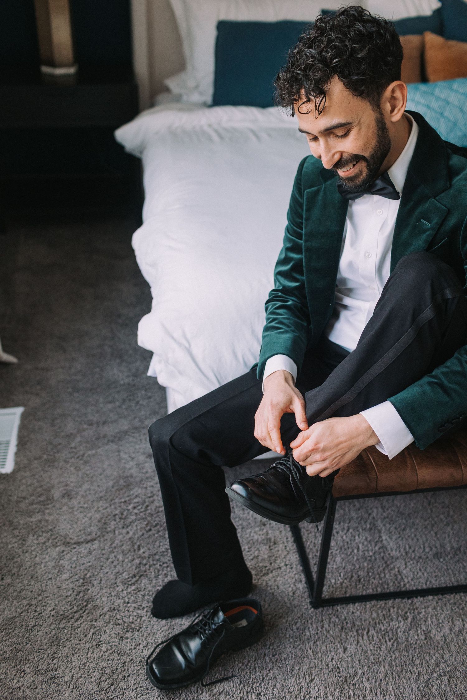 Person in a dark green suit and white shirt adjusts their shoes while sitting on a gray carpet.