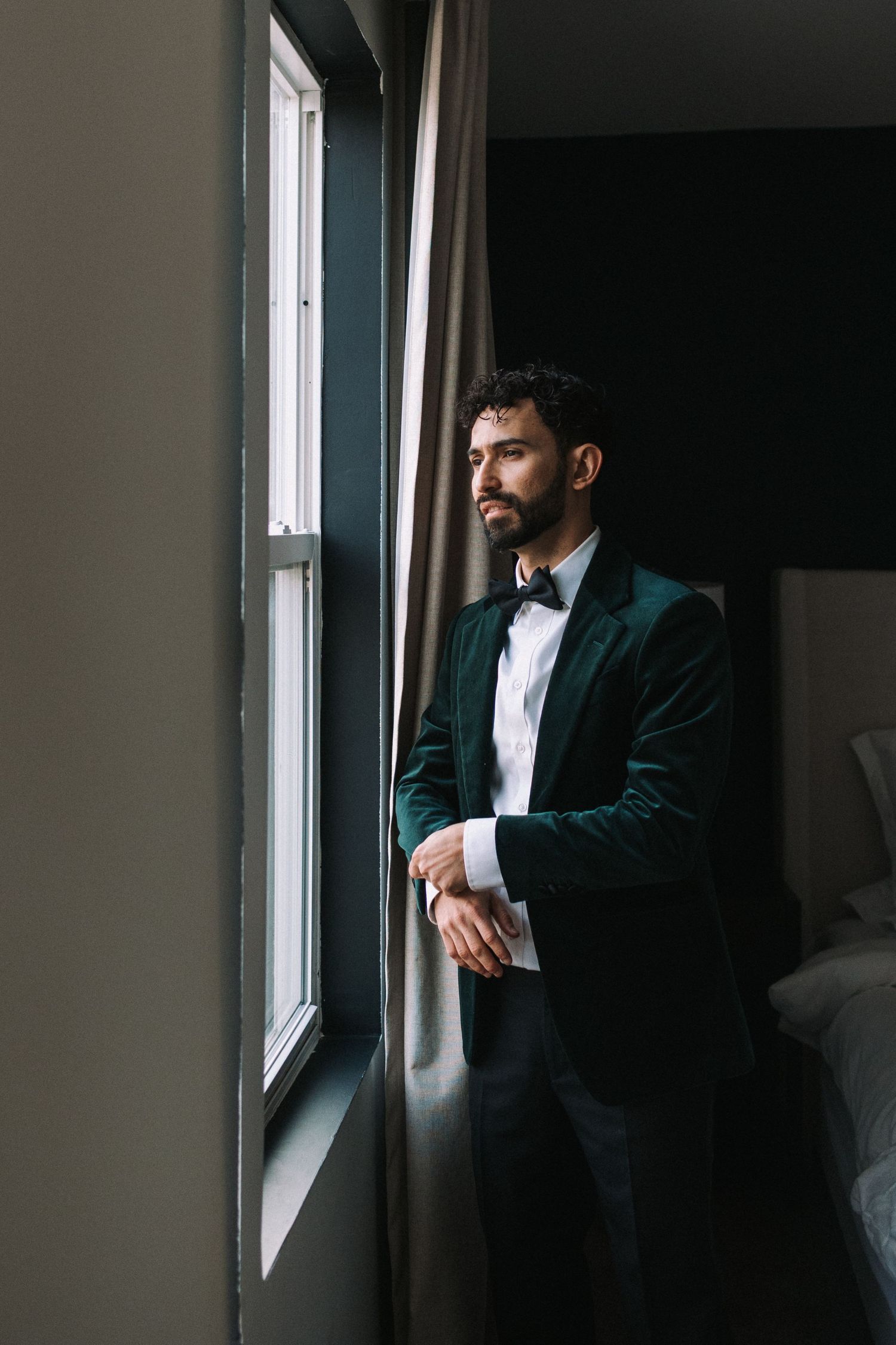 Man in dark green velvet blazer and white shirt gazes contemplatively out of a window in moody lighting.