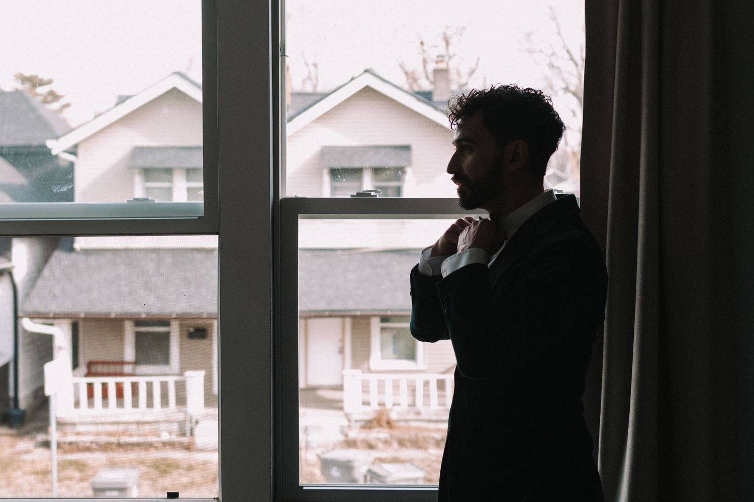 A silhouetted figure stands by a window looking out at suburban houses on a winter day.