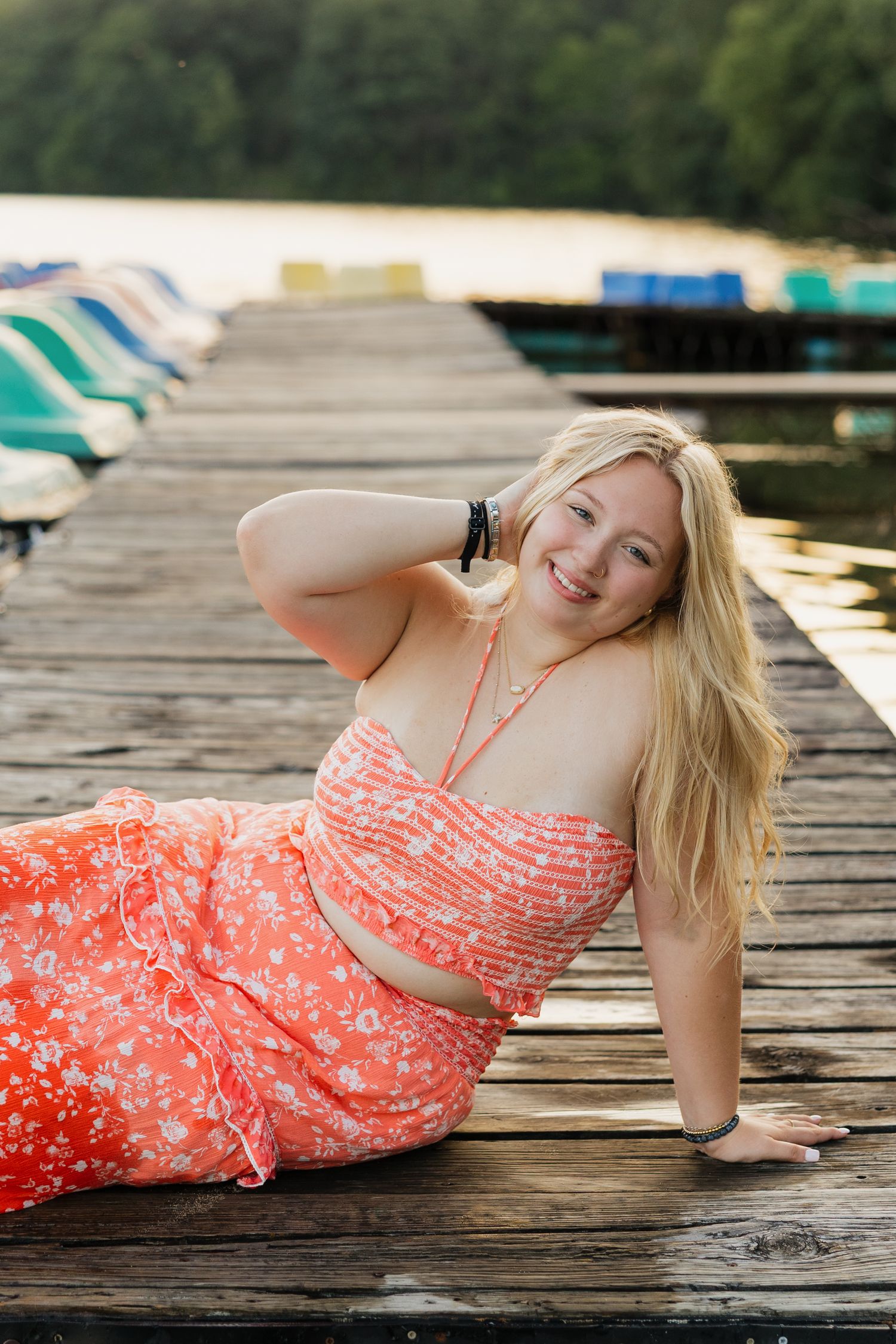 A person in a coral floral dress relaxes on a wooden dock during golden hour.