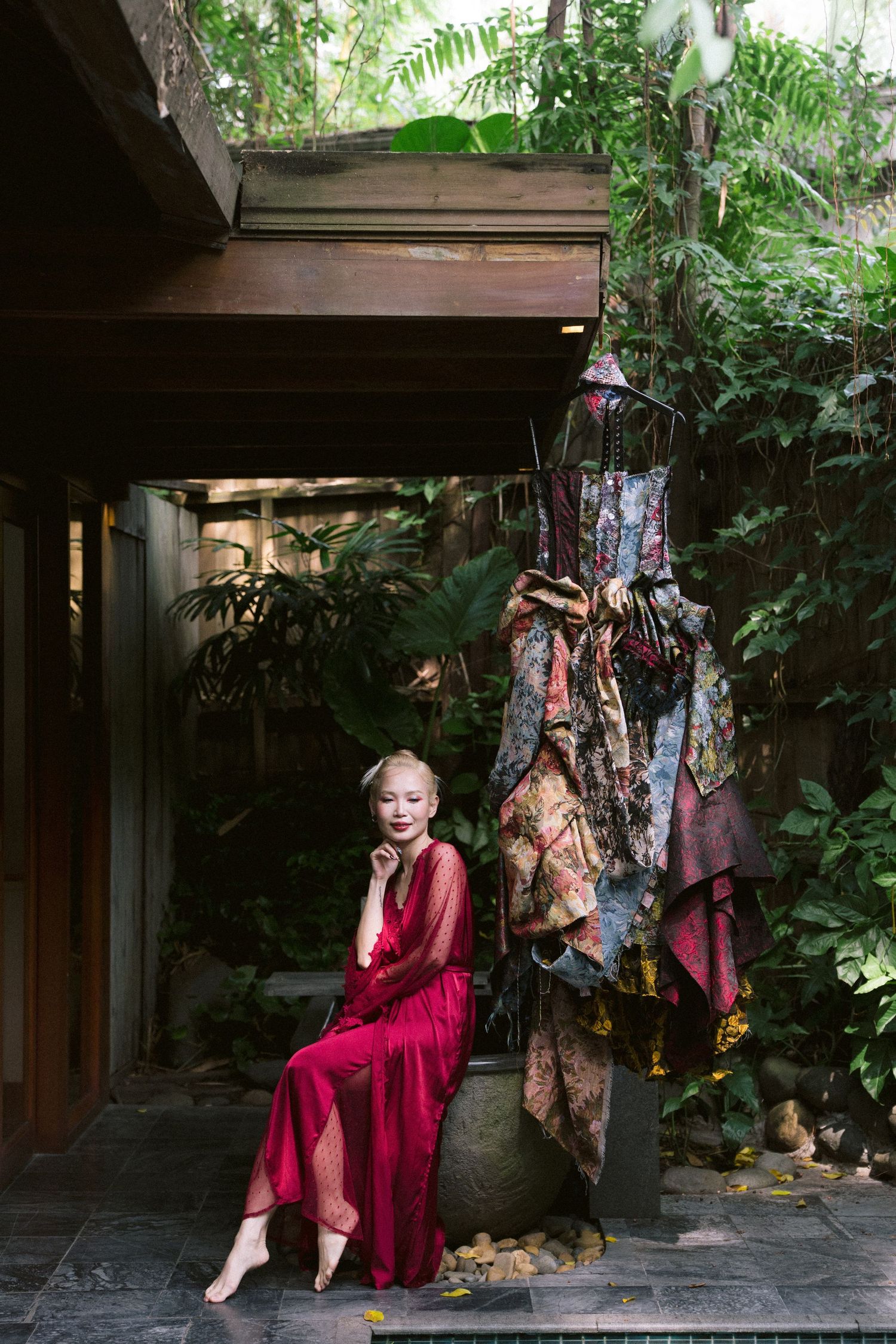 A person in a flowing red dress sits on a wooden porch surrounded by lush greenery and decorative hanging elements.