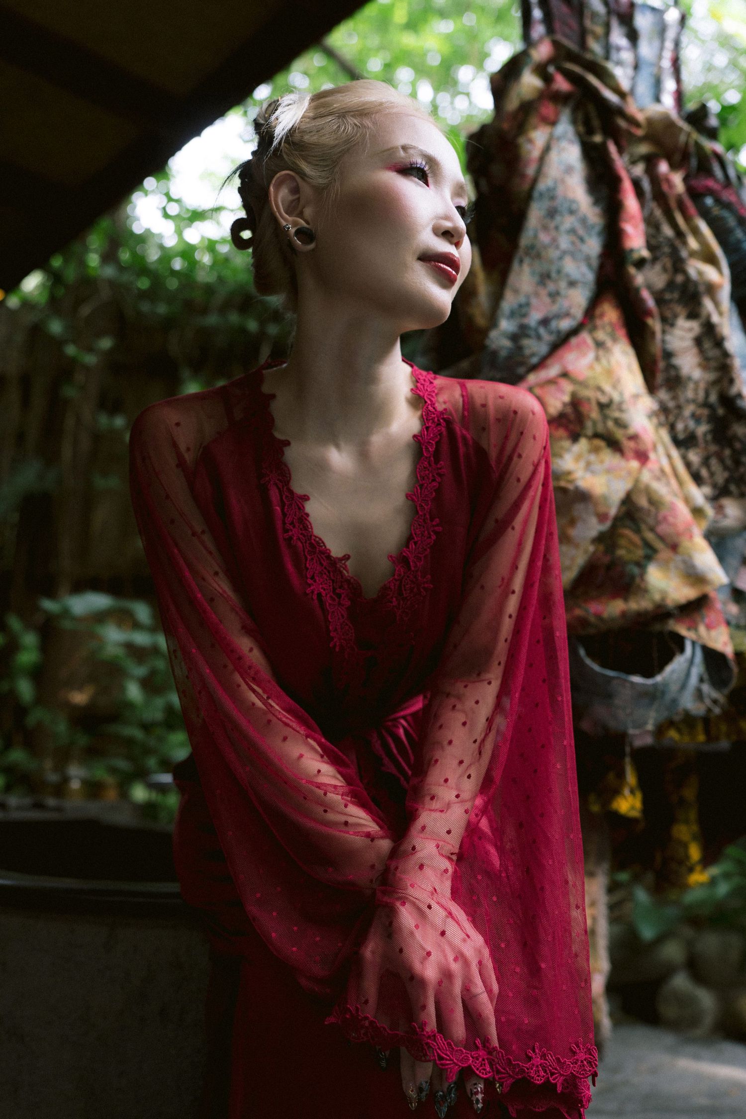A person in a flowing red lace dress poses thoughtfully against a natural outdoor backdrop.