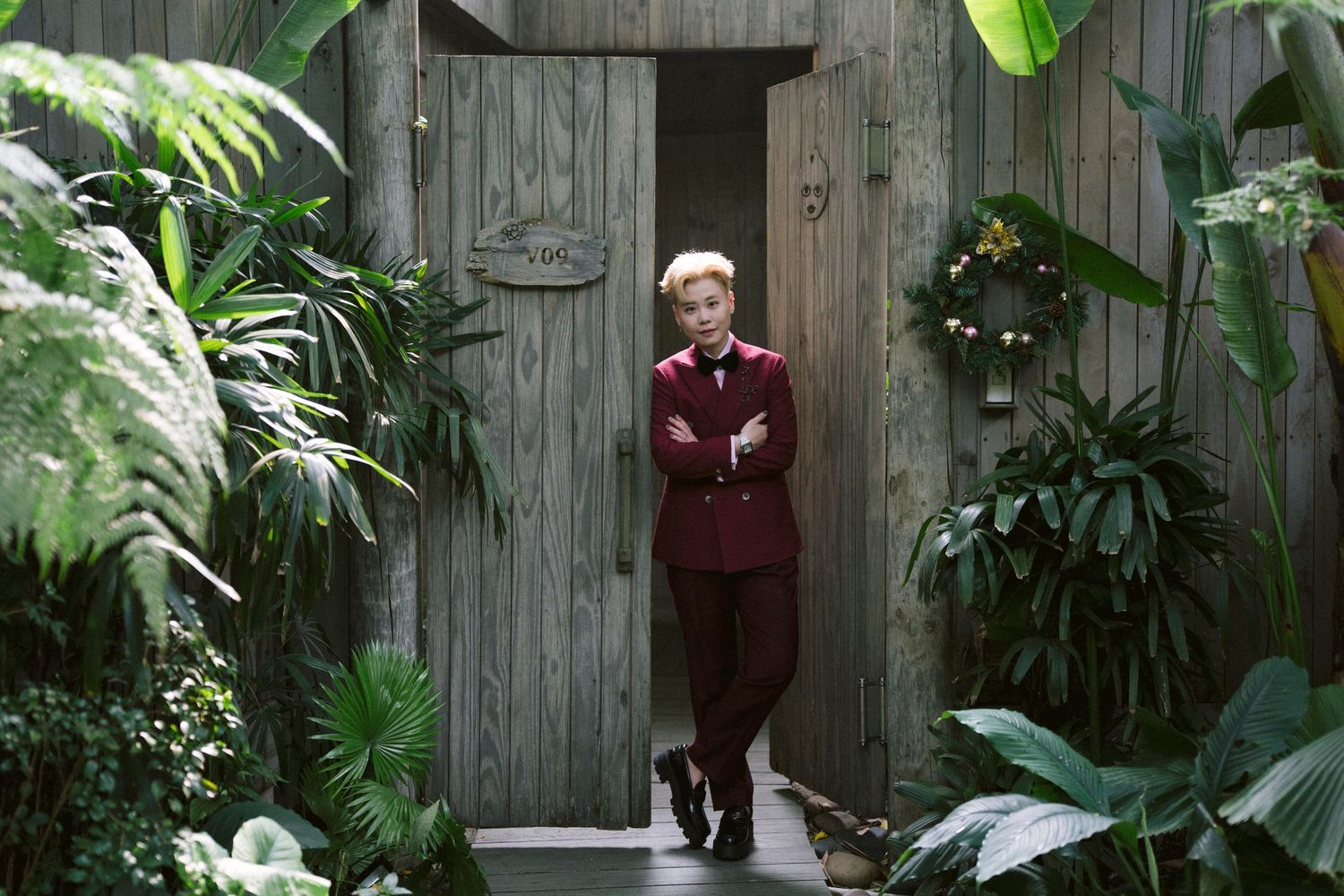A figure stands in front of a rustic wooden door surrounded by lush tropical plants and greenery.