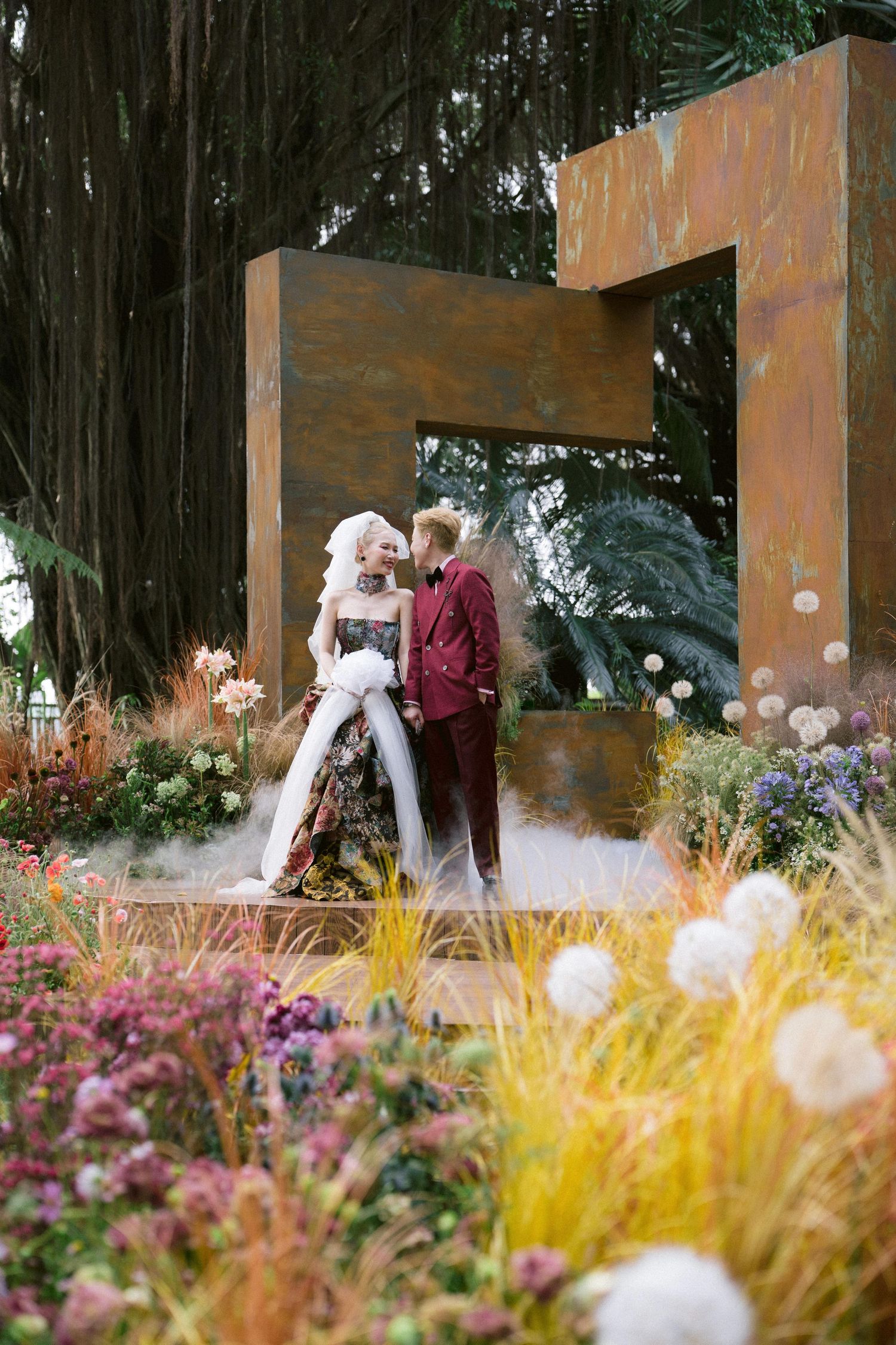 A romantic garden wedding photo featuring a couple posing in front of a rustic metal archway surrounded by colorful flowers.