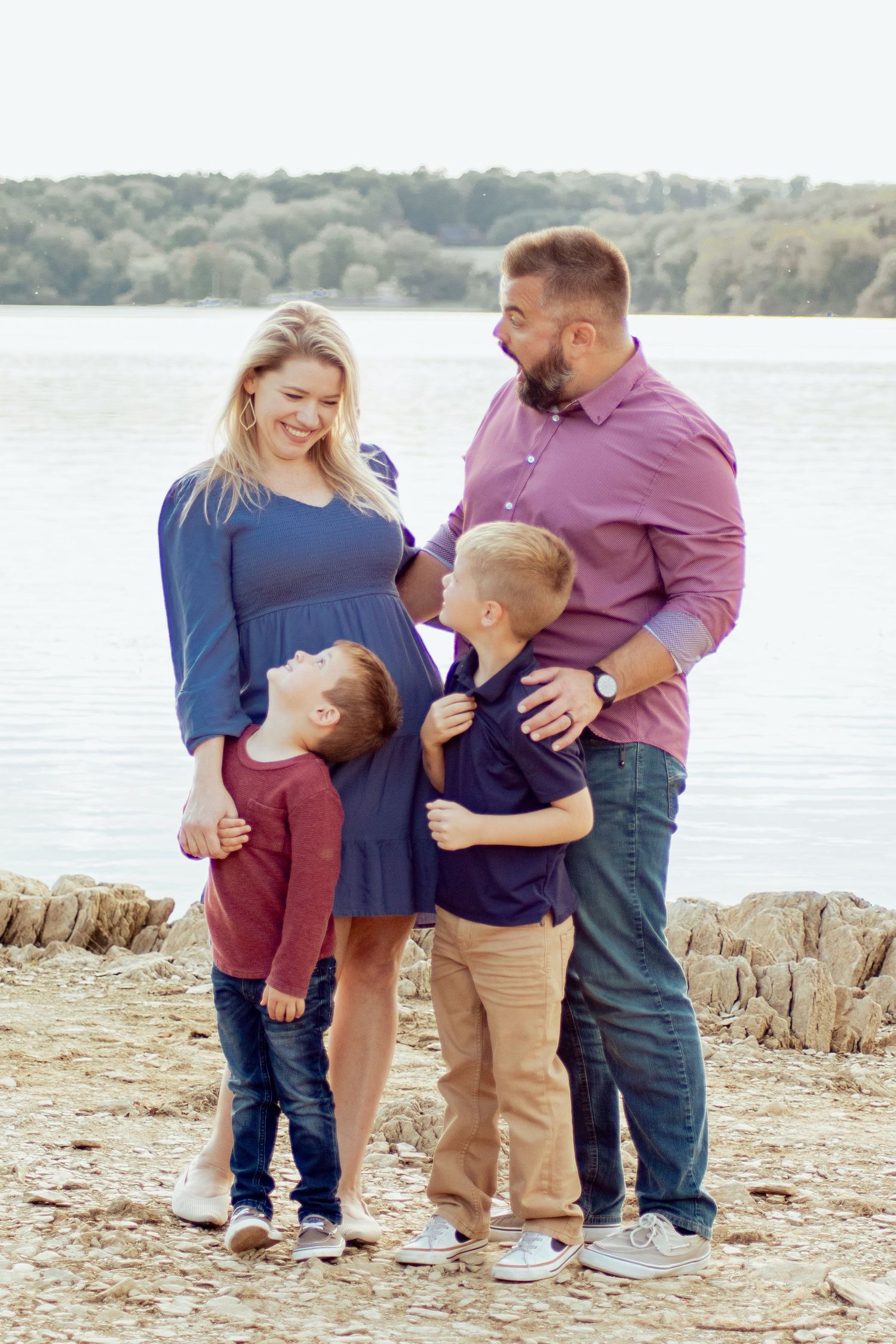 Happy pregnant family with two young boys hugging moms baby bump by a scenic lakeside shore.