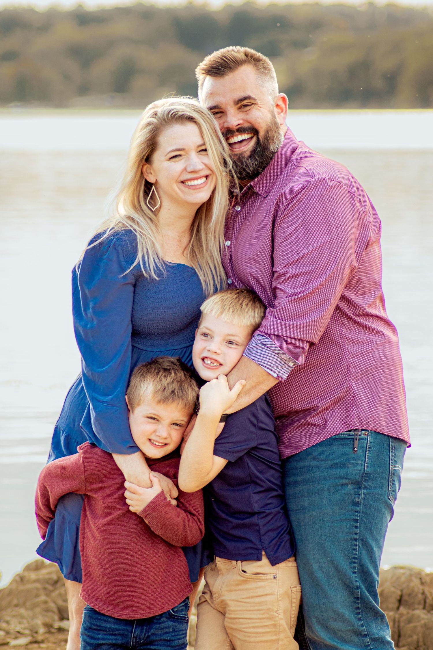Happy family of four poses by a lake, parents smiling with two young boys in coordinated blue and burgundy outfits.