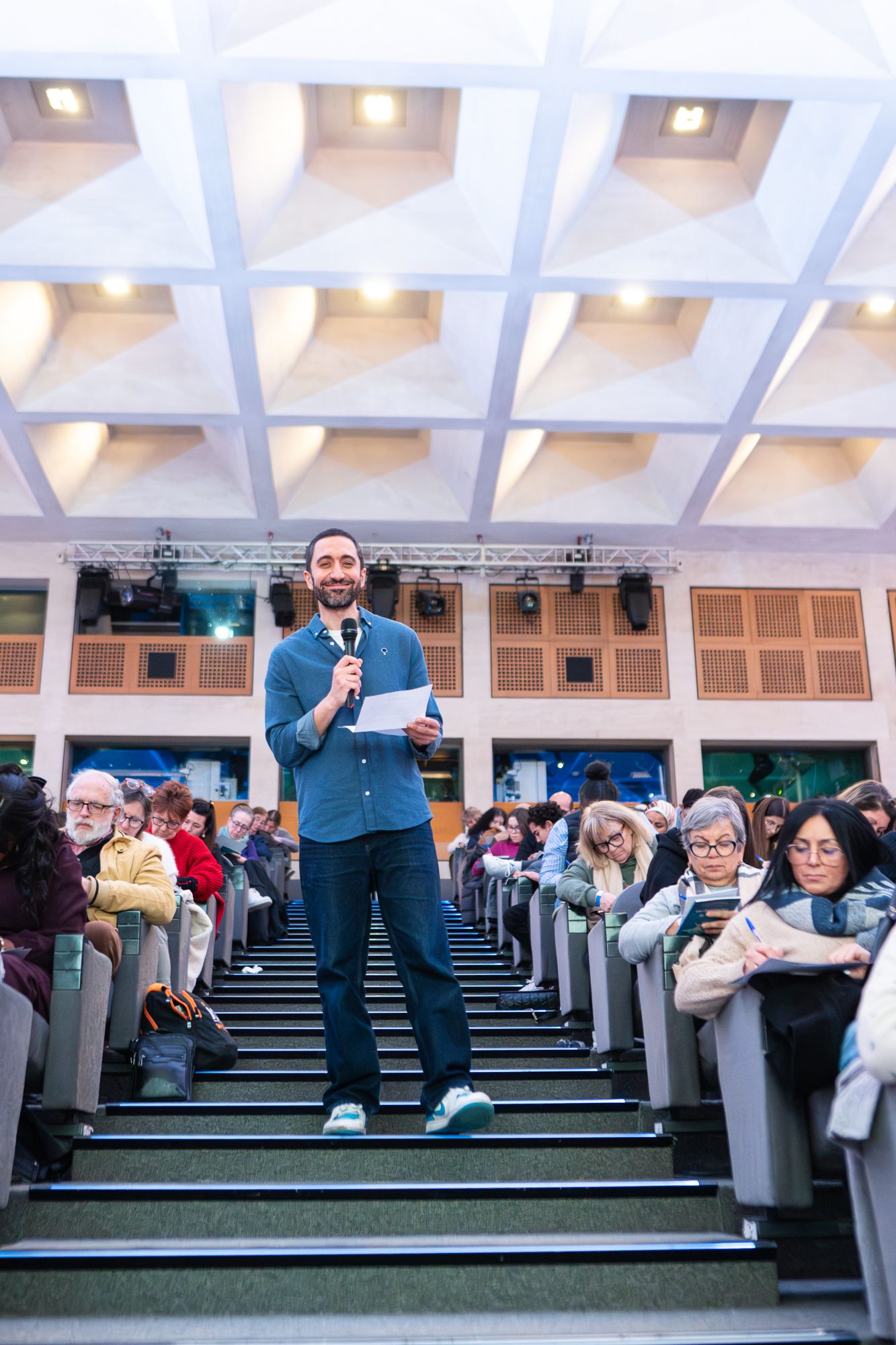A speaker stands on tiered seating while addressing an audience seated in an auditorium with geometric ceiling design.