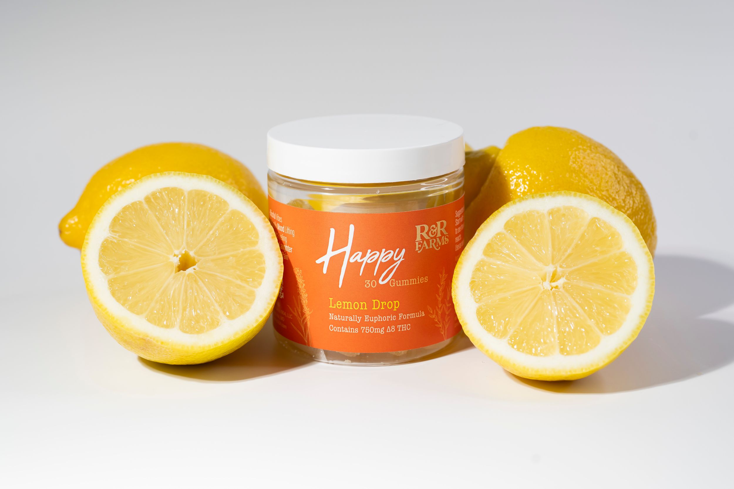 R&R Farms Happy Lemon Drop CBD gummies jar surrounded by fresh cut lemons on white background.