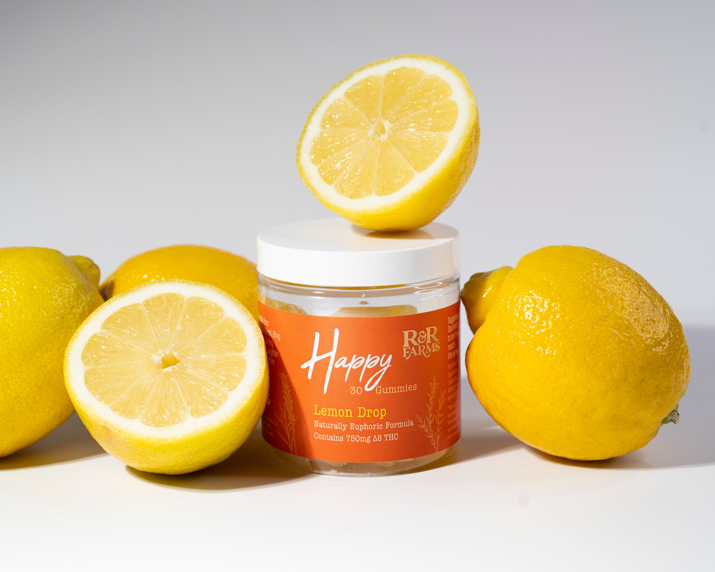 R&R Farms Happy Lemon Drop CBD gummies jar with fresh lemons on a light gray background.
