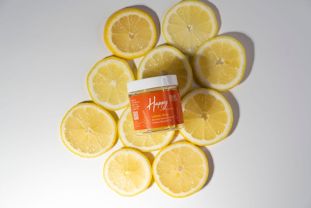 R&R Farms Happy Lemon Drop CBD gummies jar surrounded by sliced lemons on white background.