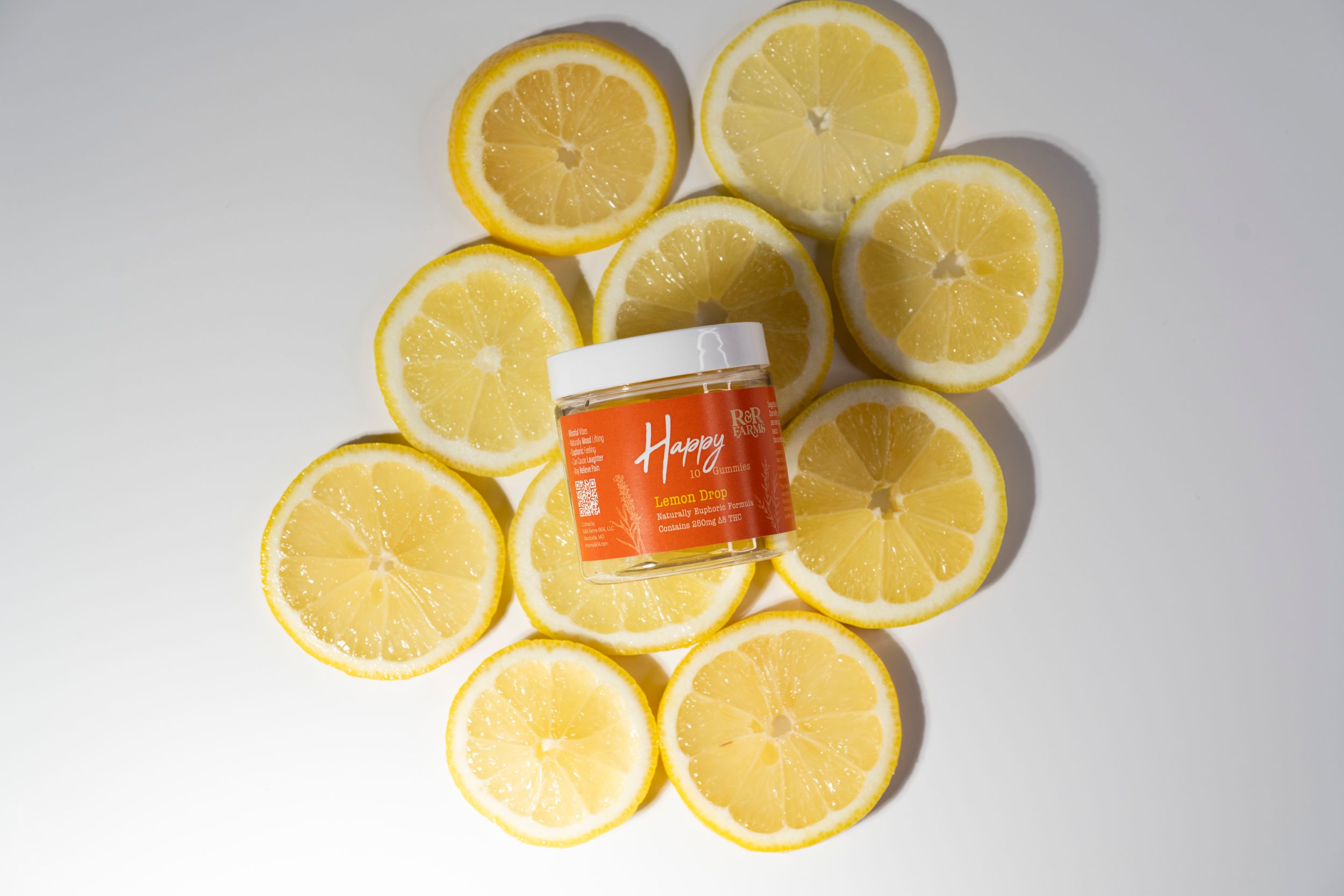 R&R Farms Happy Lemon Drop CBD gummies jar surrounded by sliced lemons on white background.