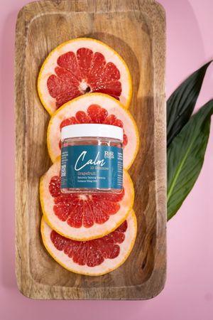 RnR Farms Calm CBD grapefruit gummies jar resting on sliced grapefruit in a wooden tray with green leaf on pink backdrop.