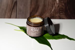 Open amber jar of Relief CBD balm with arnica and cannabis flower resting on a tropical green leaf on white surface.