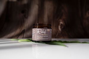 Amber glass jar of Relief CBD balm with arnica and cannabis flower placed on green leaf against dark wood background.