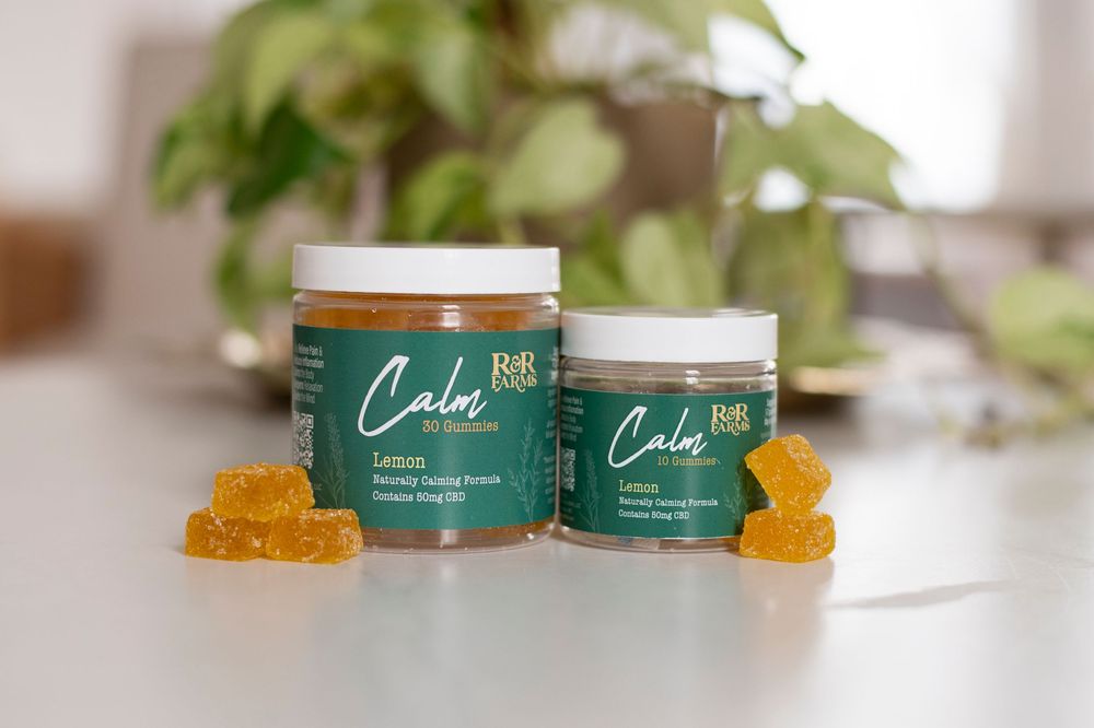 Two RnR Farms Calm CBD lemon gummies jars in different sizes displayed with golden gummies on countertop near houseplant.