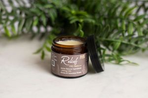 Open amber jar of RnR Farms Relief CBD balm with arnica and cannabis flower on green leaf with evergreen background.