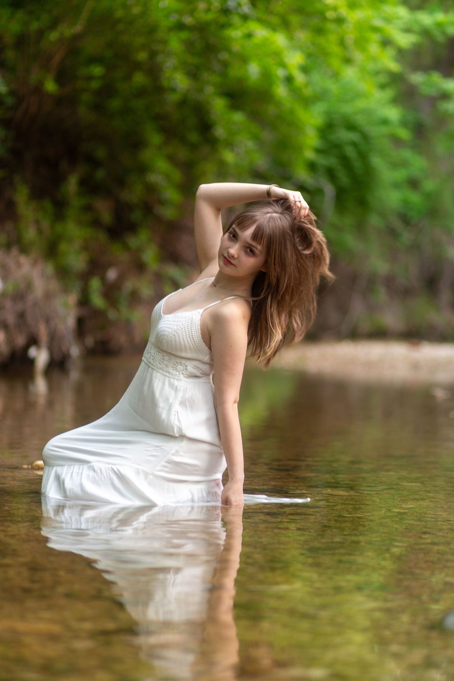 A figure in a flowing white dress poses gracefully in shallow water surrounded by lush greenery. Senior session Airfield Falls Fort Worth TX