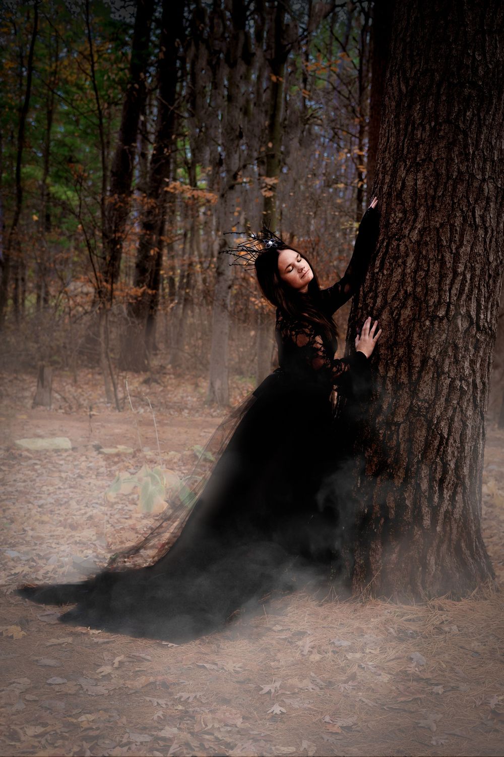 A person in a flowing black dress poses dramatically against a tree trunk in a misty forest setting.