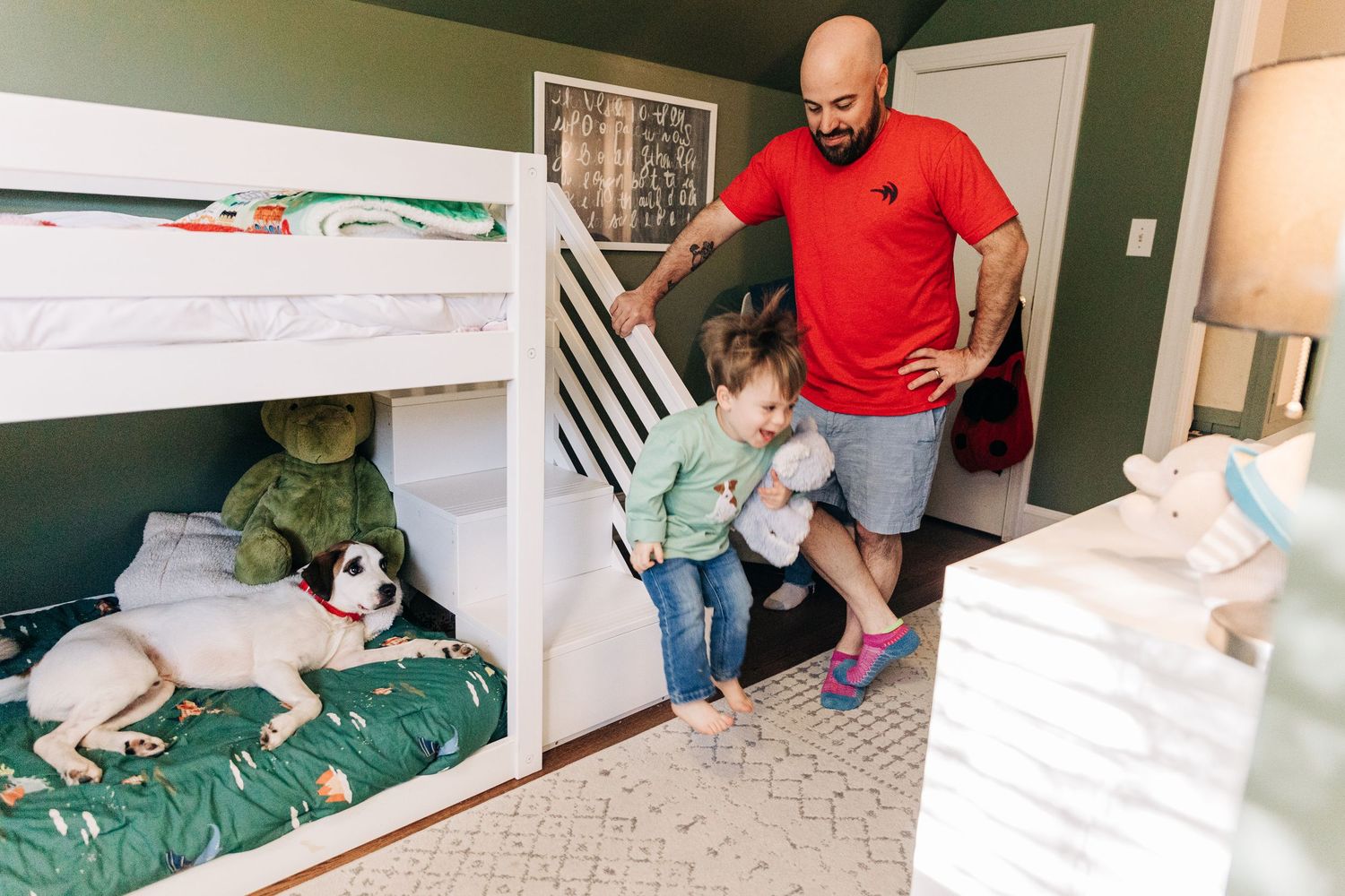 A parent watches a small child jump down from a bunk bed while a dog rests on the bottom bunk in a cozy bedroom.