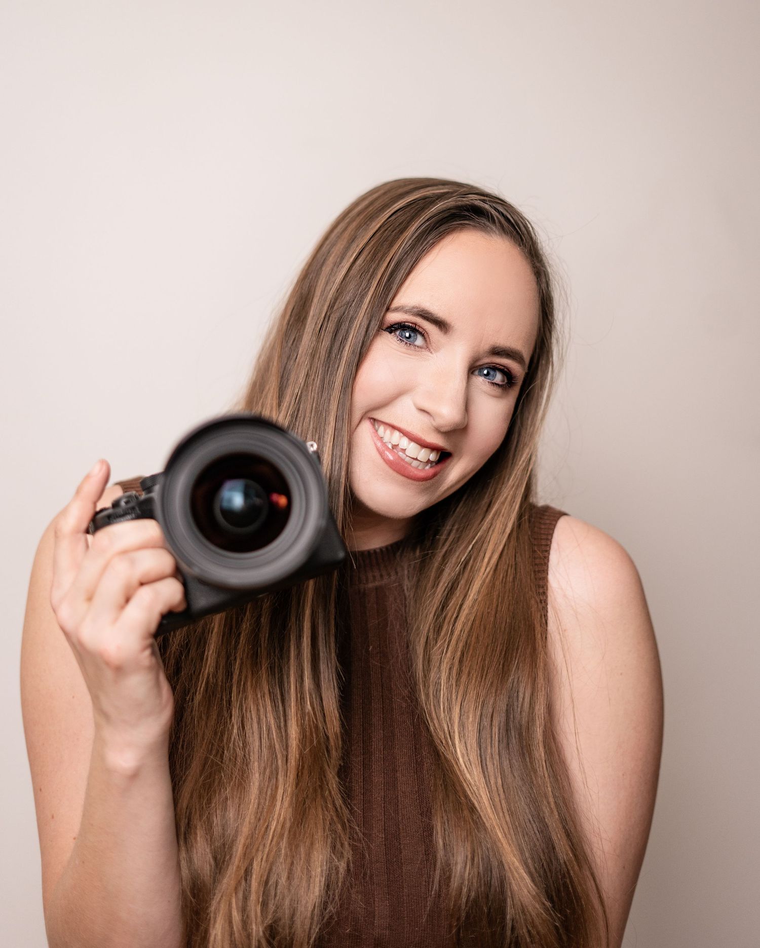 A photographer with long brown hair smiles while holding a professional camera lens against a neutral background.