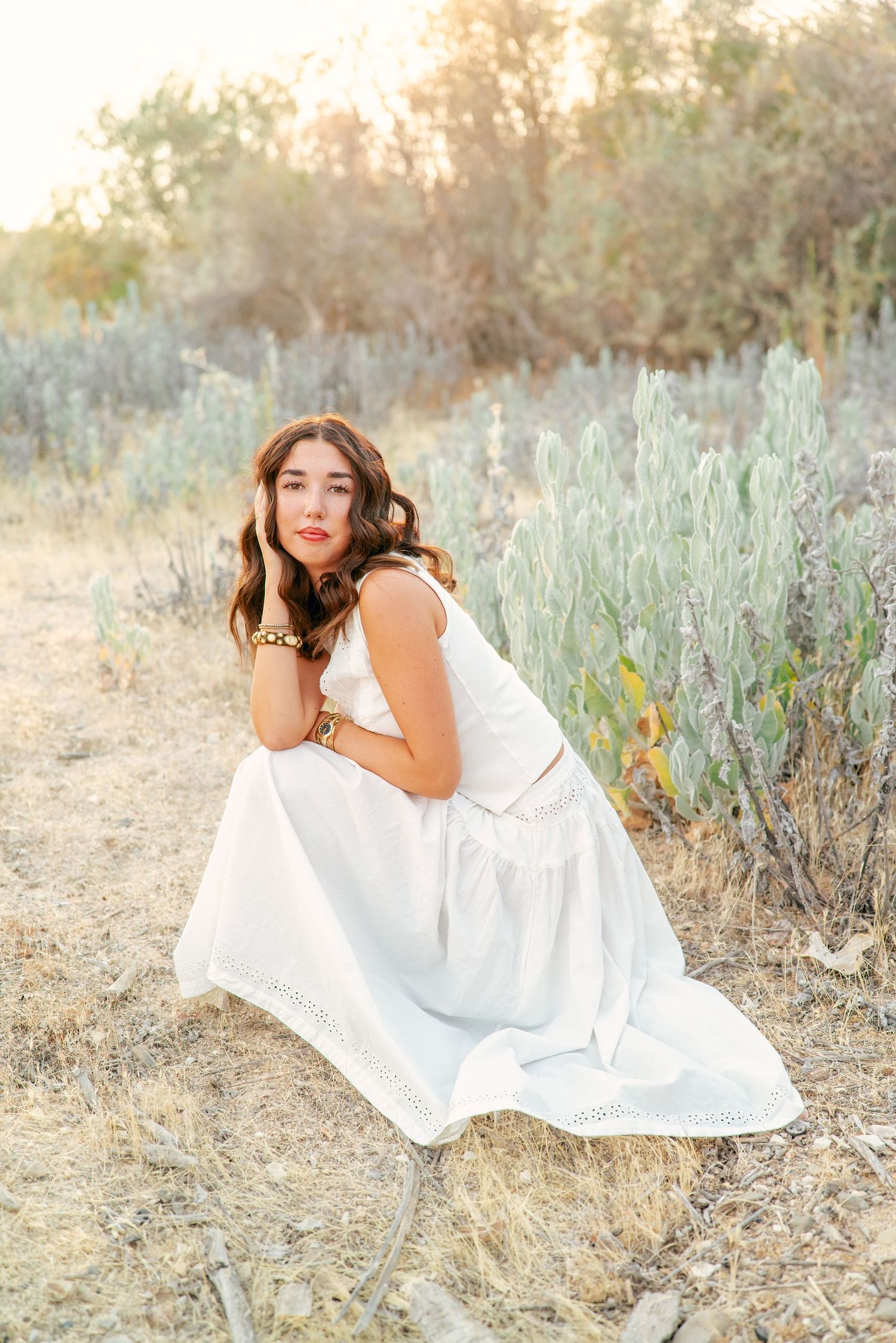 A woman in a flowing white dress sits gracefully on the ground surrounded by sage brush at sunset.