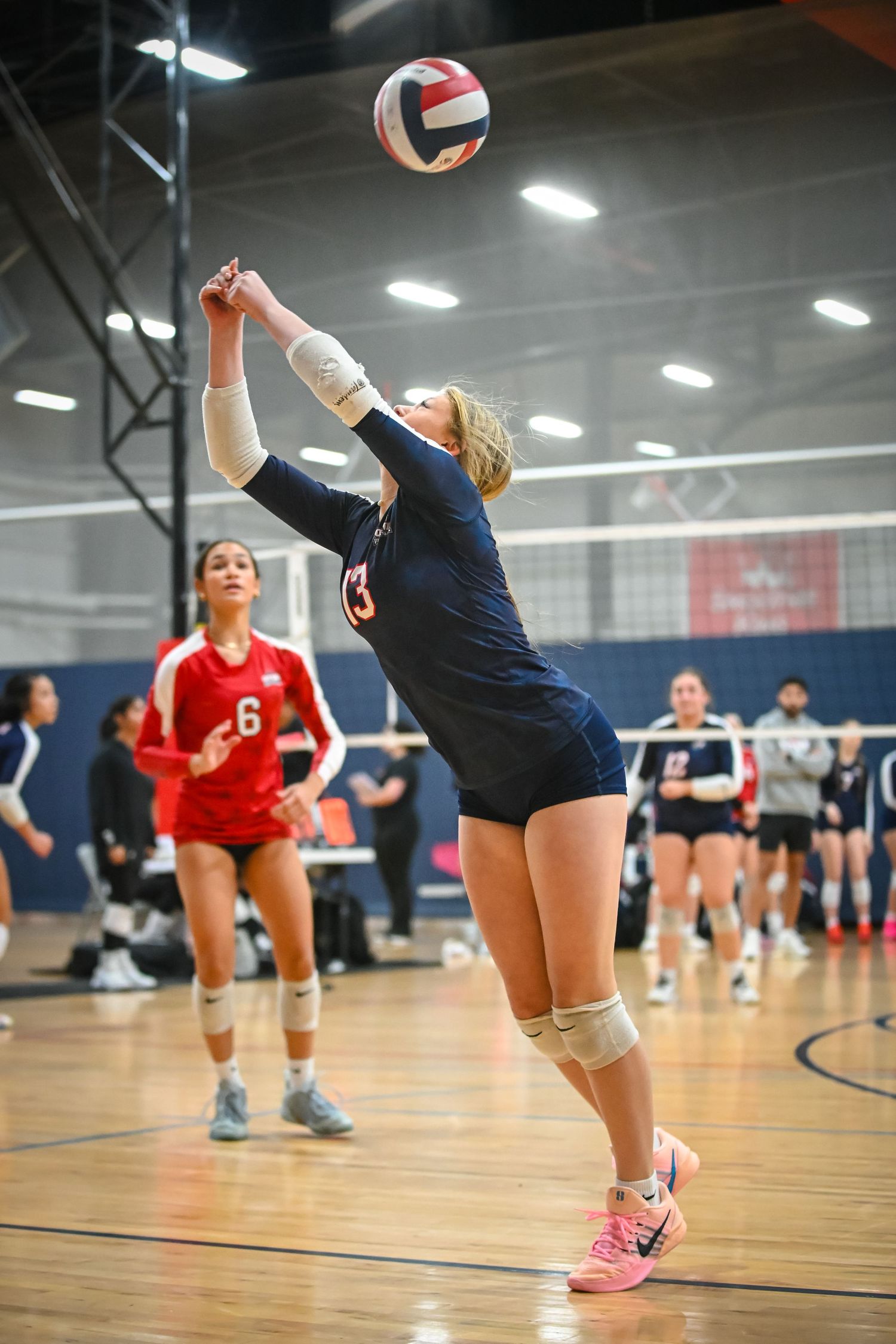 Indoor volleyball players compete in a recreational match on a wooden court with red and navy uniforms.