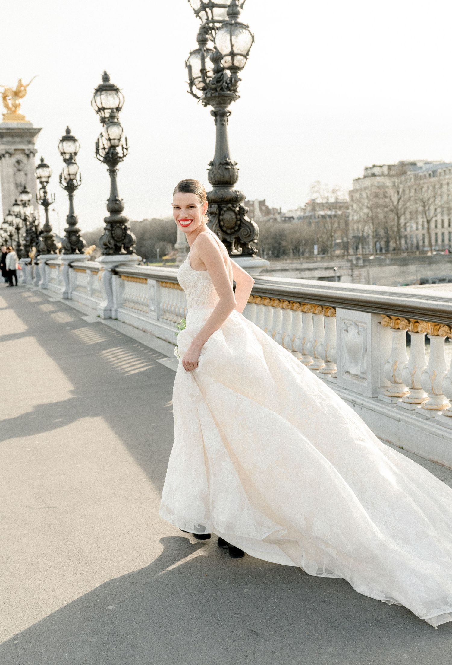 A bride in a flowing white gown poses gracefully on a historic bridge with ornate lampposts and Parisian architecture in view.