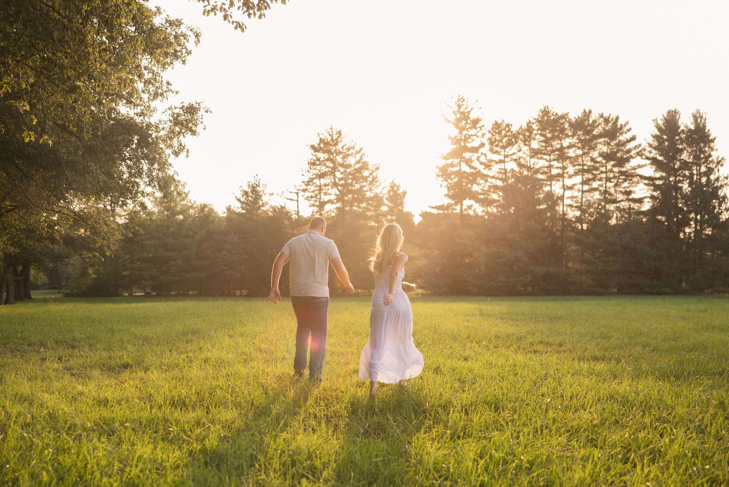 Couple walking hand in hand through a sunlit meadow at golden hour with tall grass and trees in background.