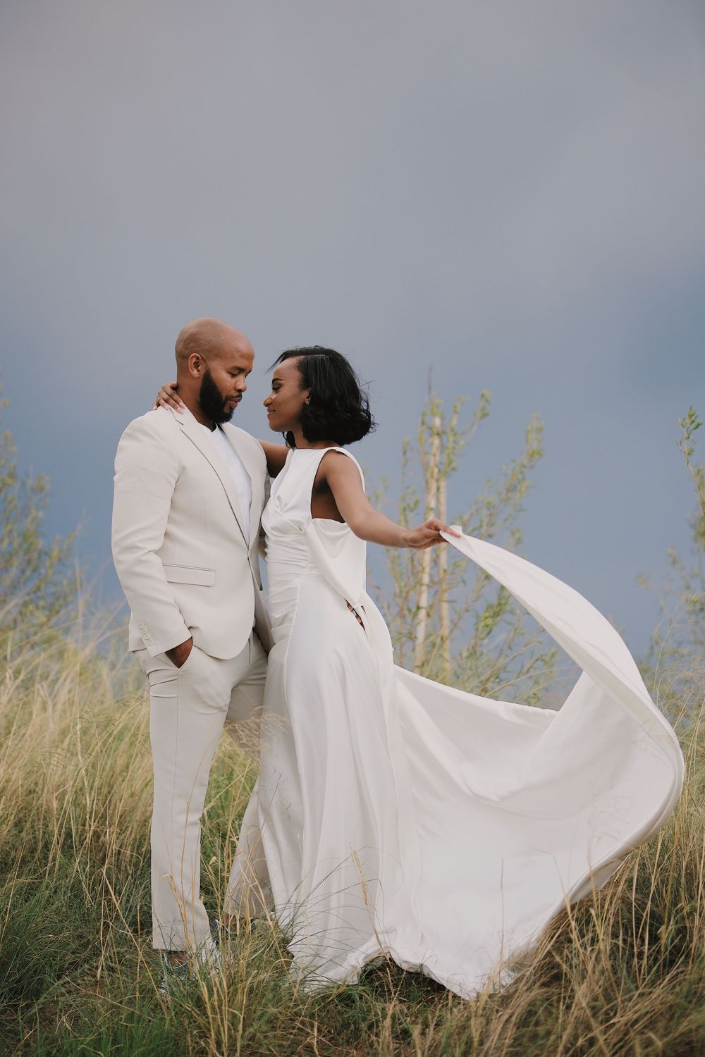 Black couple in white wedding attire embracing in tall grass field with dramatic cloudy sky.