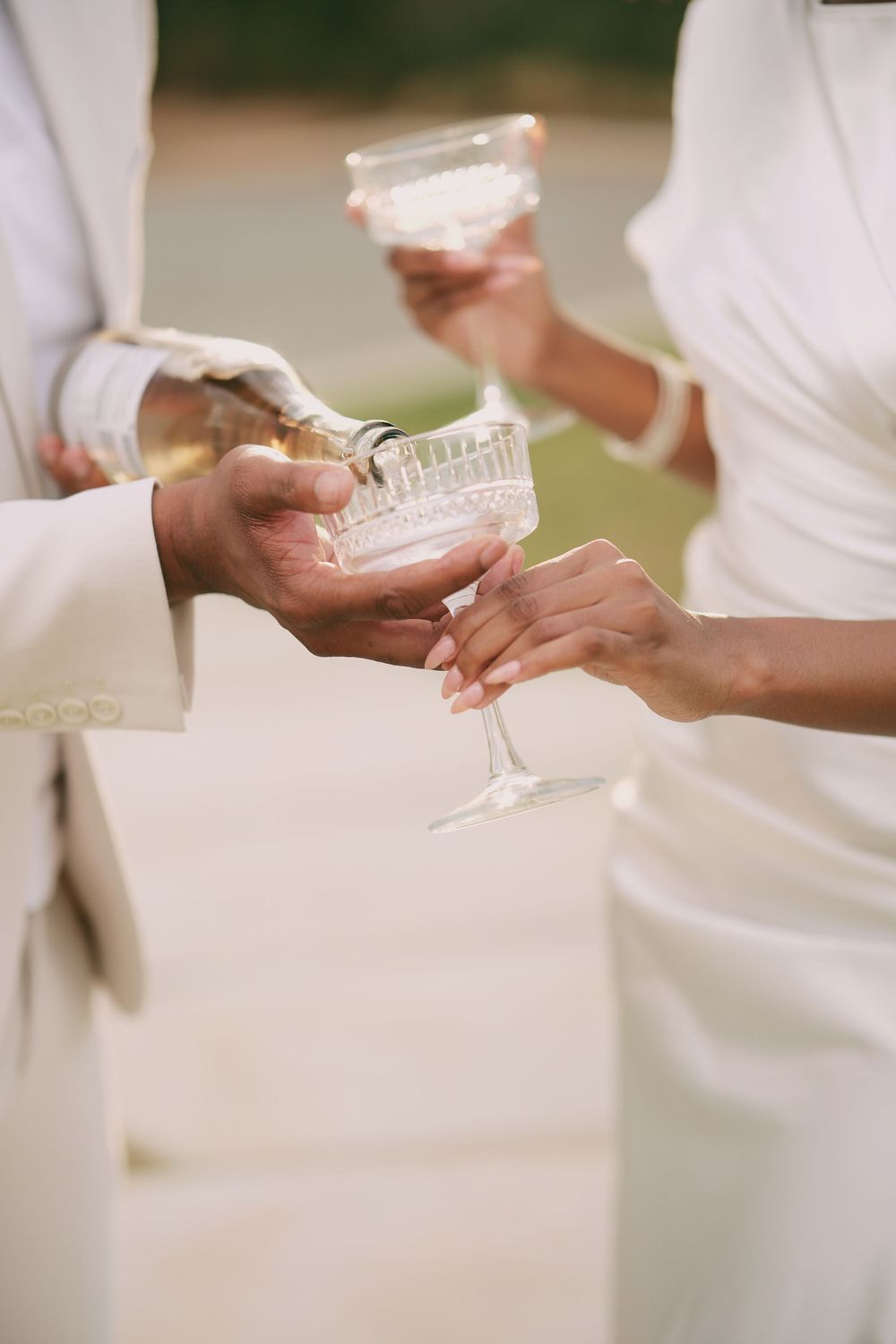 Couple in white wedding attire pouring champagne into crystal coupe glasses outdoors.