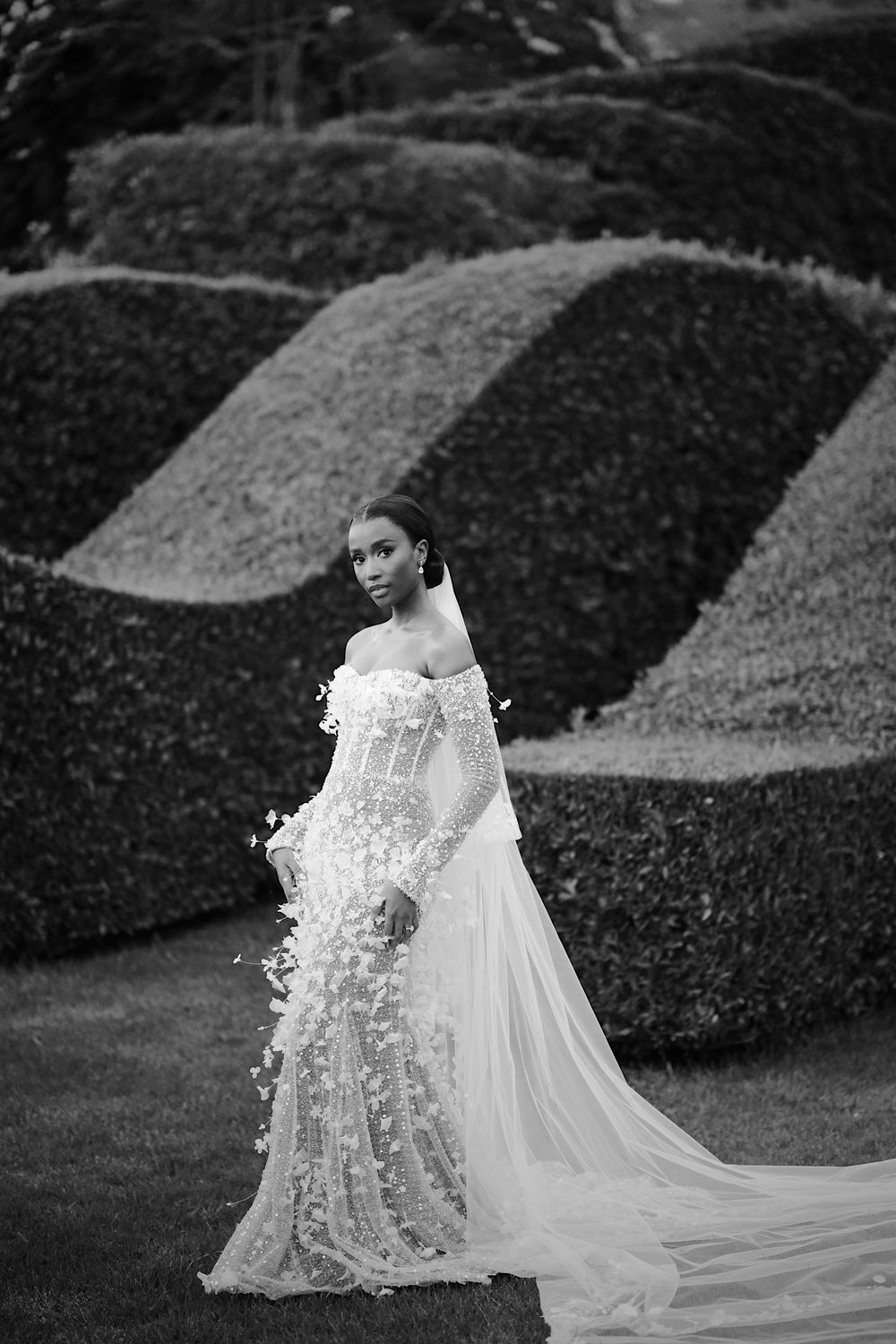 Black bride in floral applique off-shoulder wedding gown posing before sculpted topiary hedges, black and white.