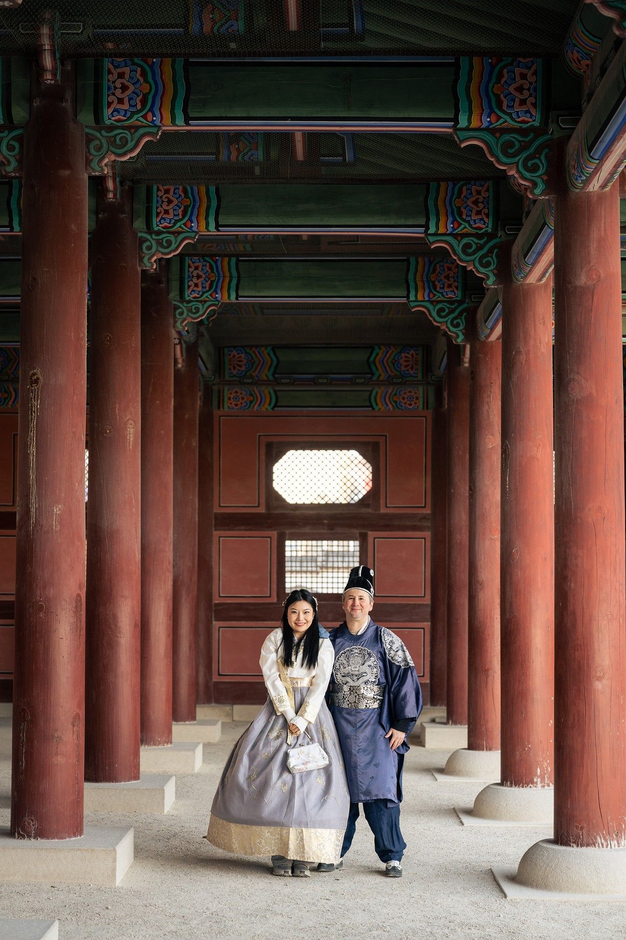 Two people stand beneath traditional red pillars in a historic Korean palace corridor.