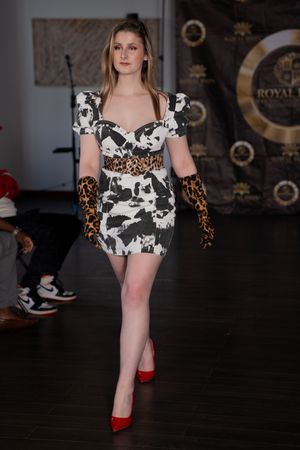 Model walks runway in black and white cow print romper with brown leopard print sleeves and red heels.