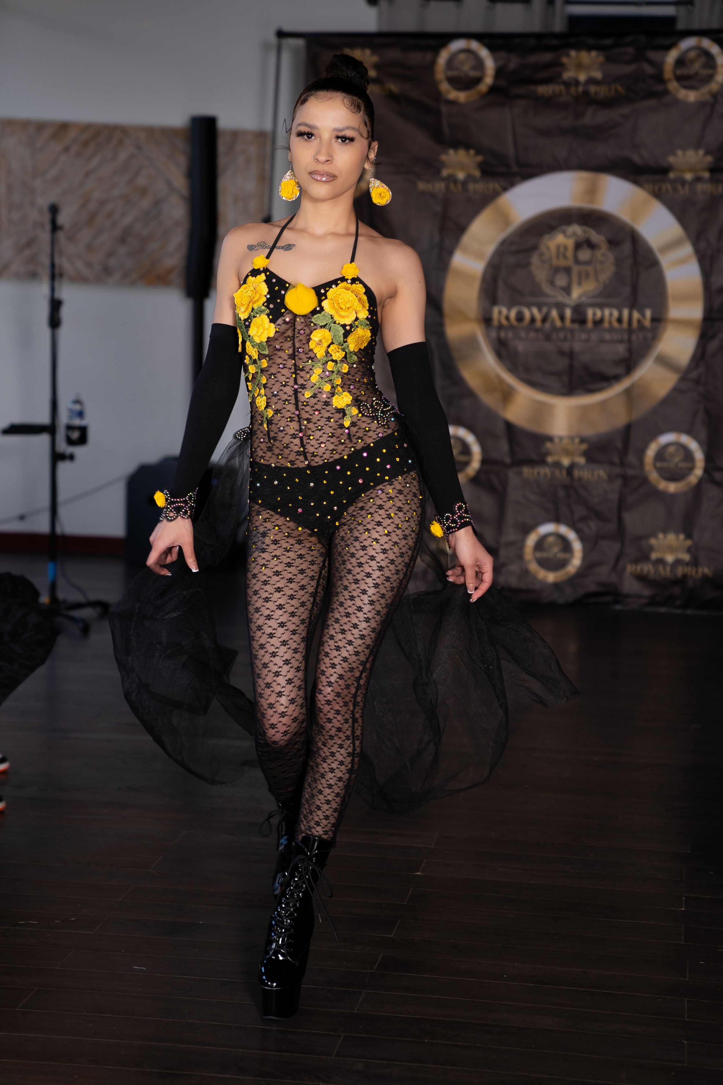 Fashion model walks runway in black lace bodysuit with yellow floral details at Royal Pride fashion show.