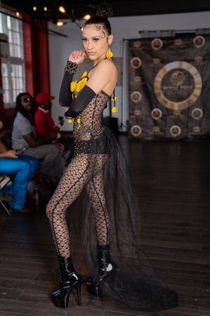 Model showcases black lace bodysuit with dramatic tulle cape on dark runway stage.