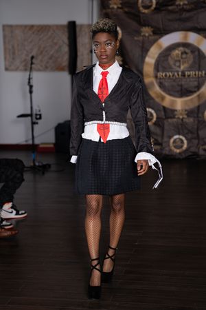A model walks the runway in a black schoolgirl-inspired outfit with white collar and red tie.