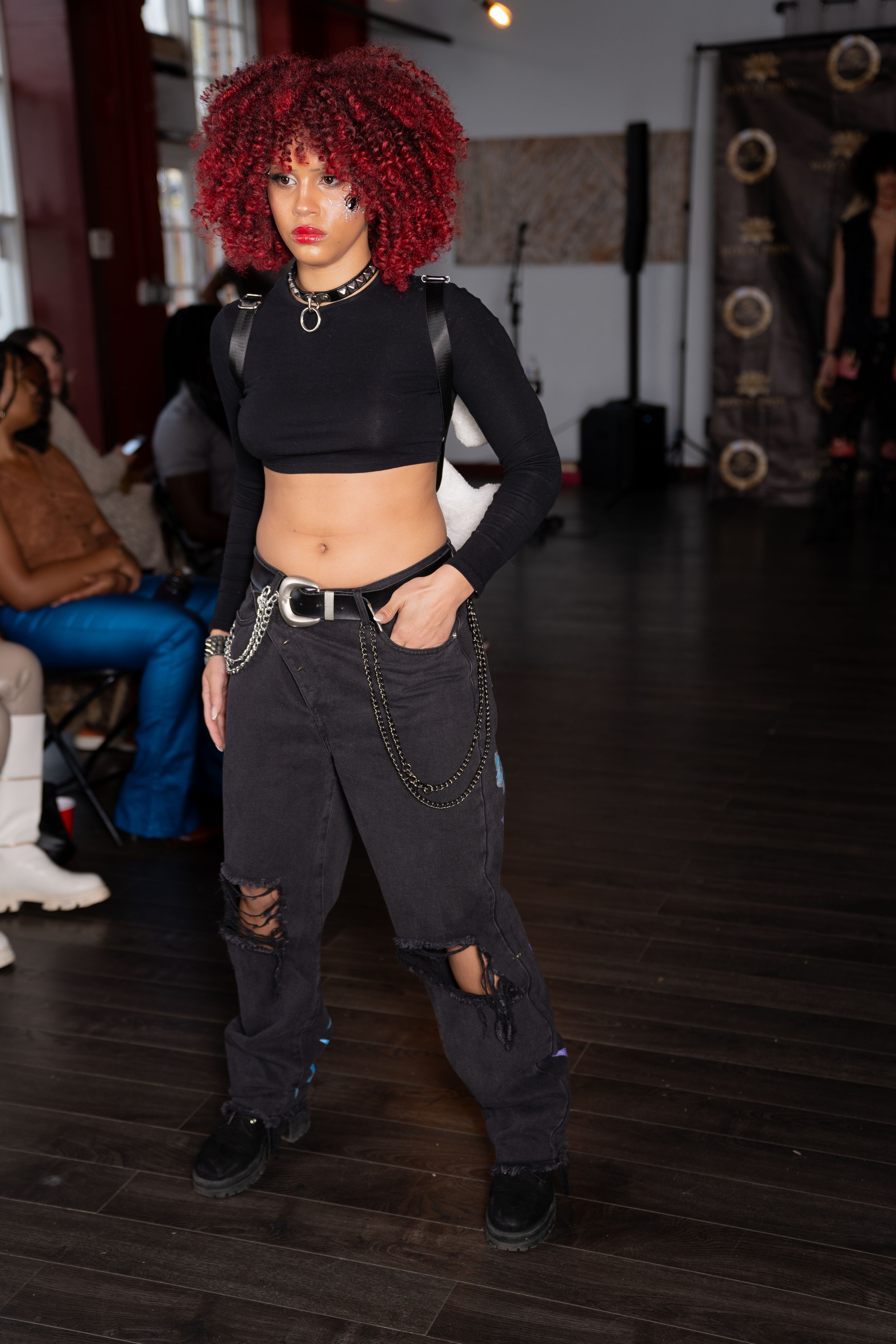 Fashion model in black streetwear ensemble and bright red hair poses on wooden floor runway.