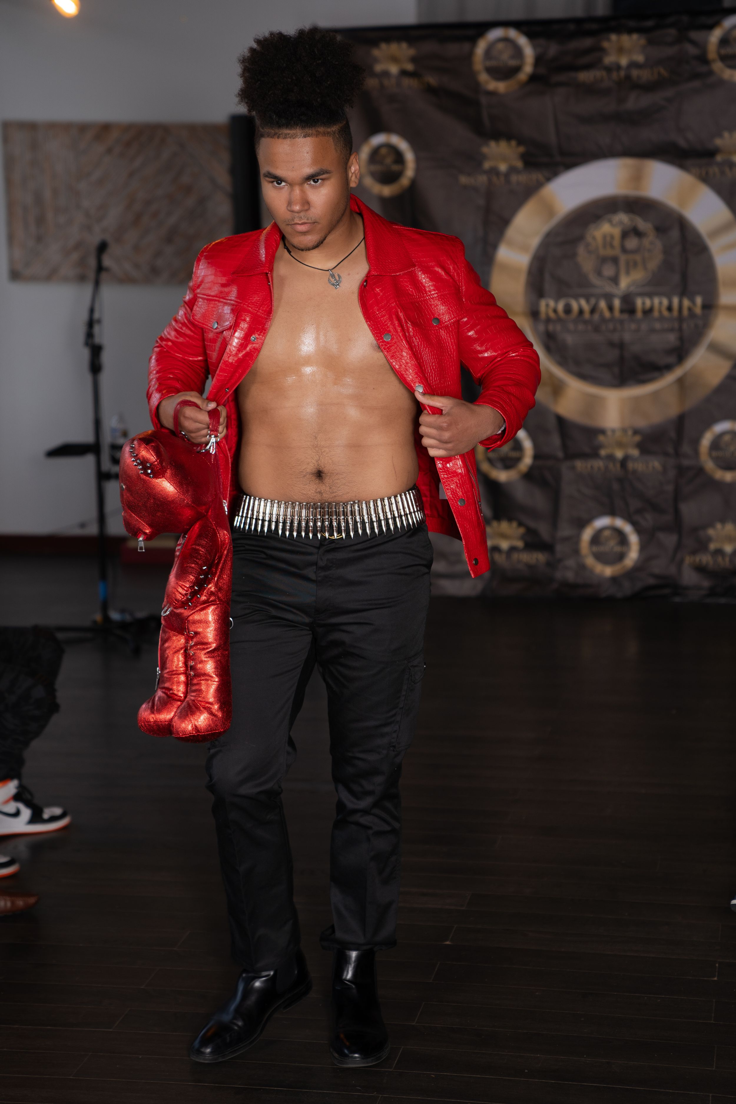 Model walks runway in red metallic jacket and black pants with silver chain belt detail.