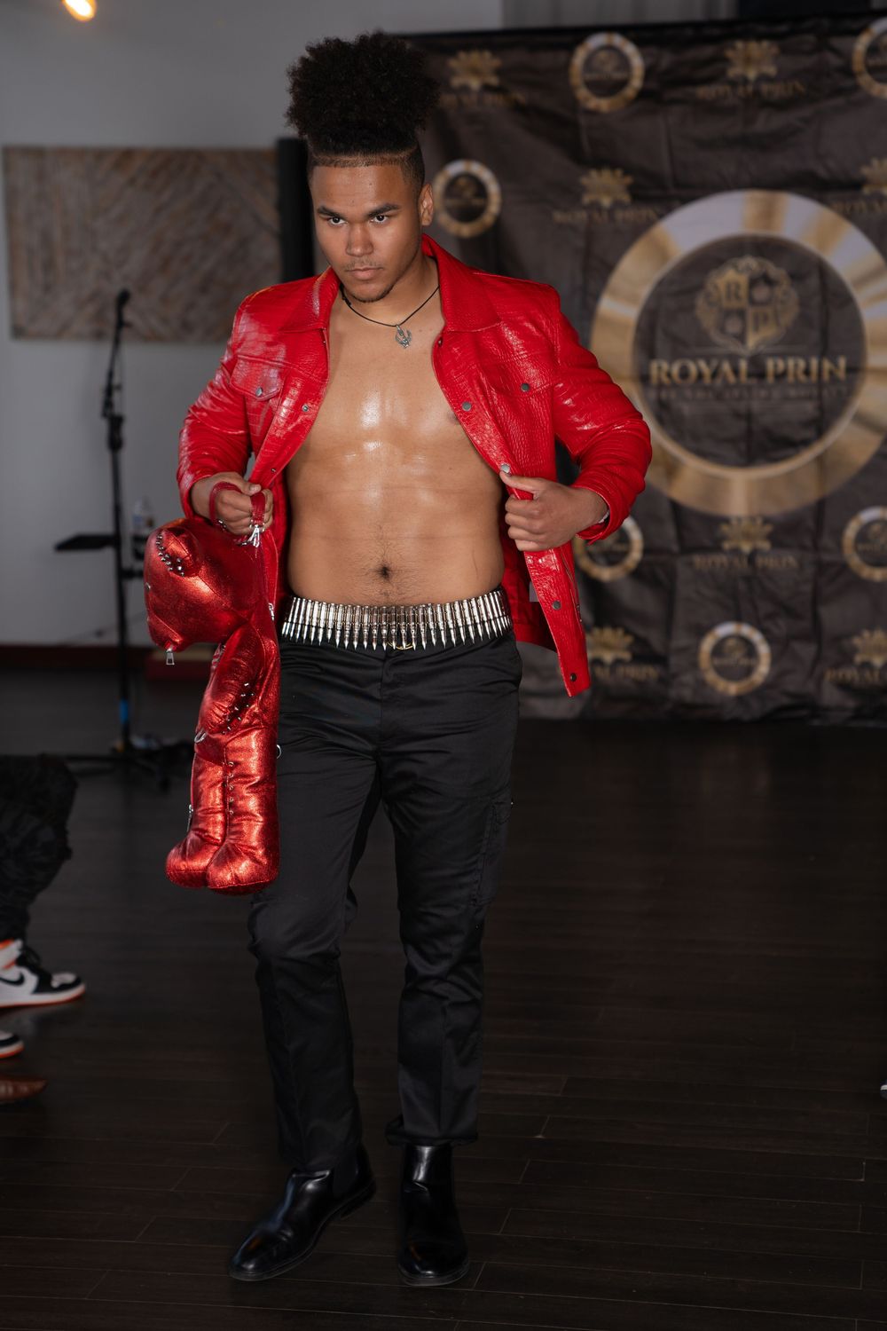 Model walks runway in red metallic jacket and black pants with silver chain belt detail.