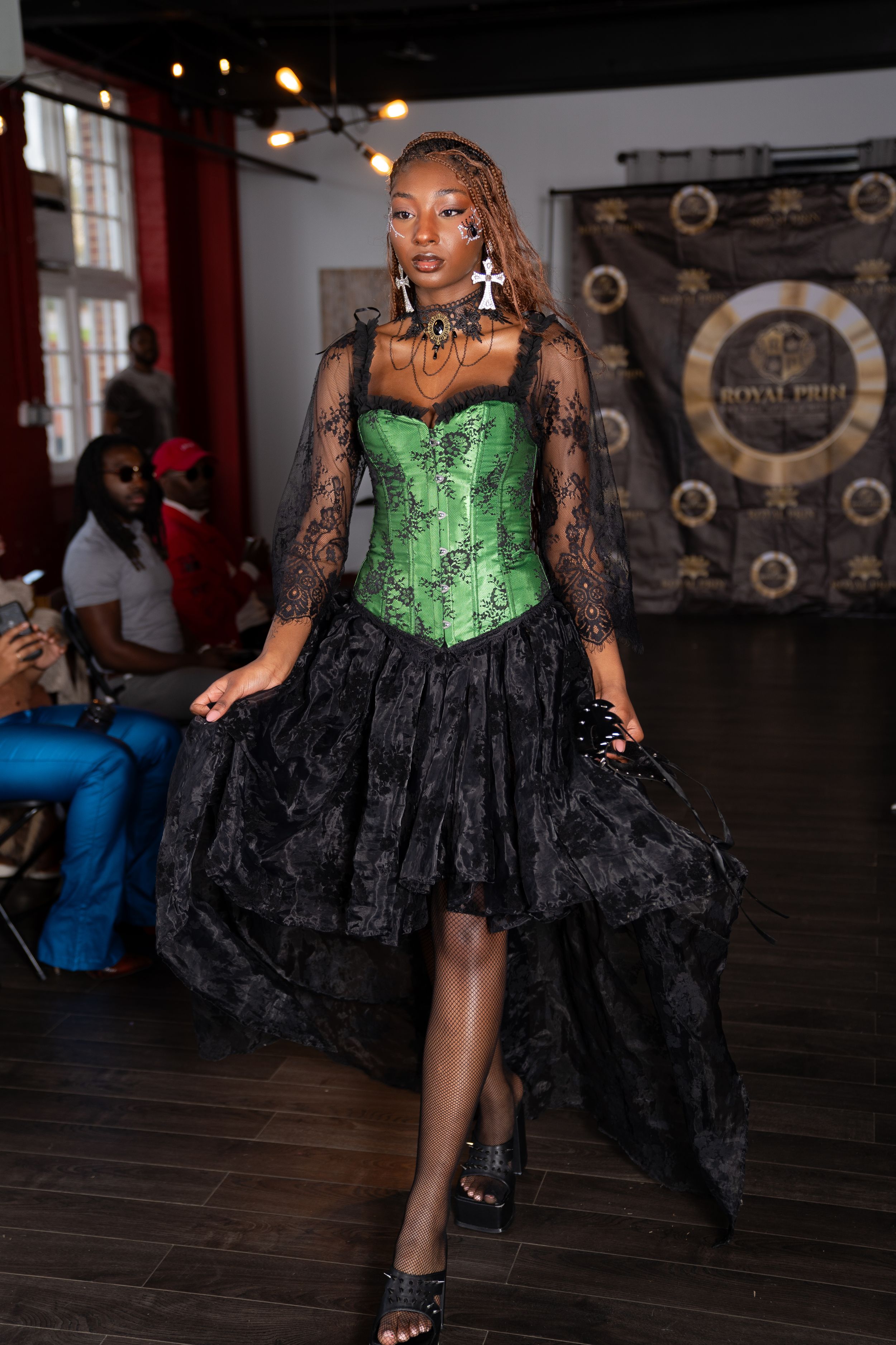 Fashion model in emerald green corset with black lace overlay and asymmetrical skirt on runway.
