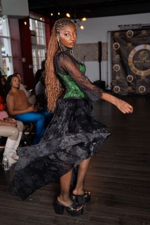A dramatic dance move in a black dress with green accents captured on a wooden stage floor.
