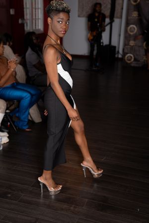 Model walks runway in elegant black evening gown with high slit and silver heels at fashion show.