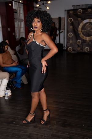A person in a fitted black dress strikes a pose on a wooden dance floor at a dimly lit venue.