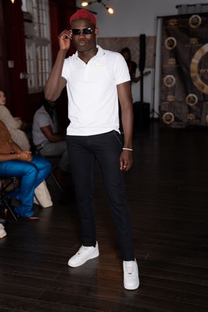 A person in a white polo shirt, dark pants and white sneakers poses on a dark wooden floor with sunglasses.