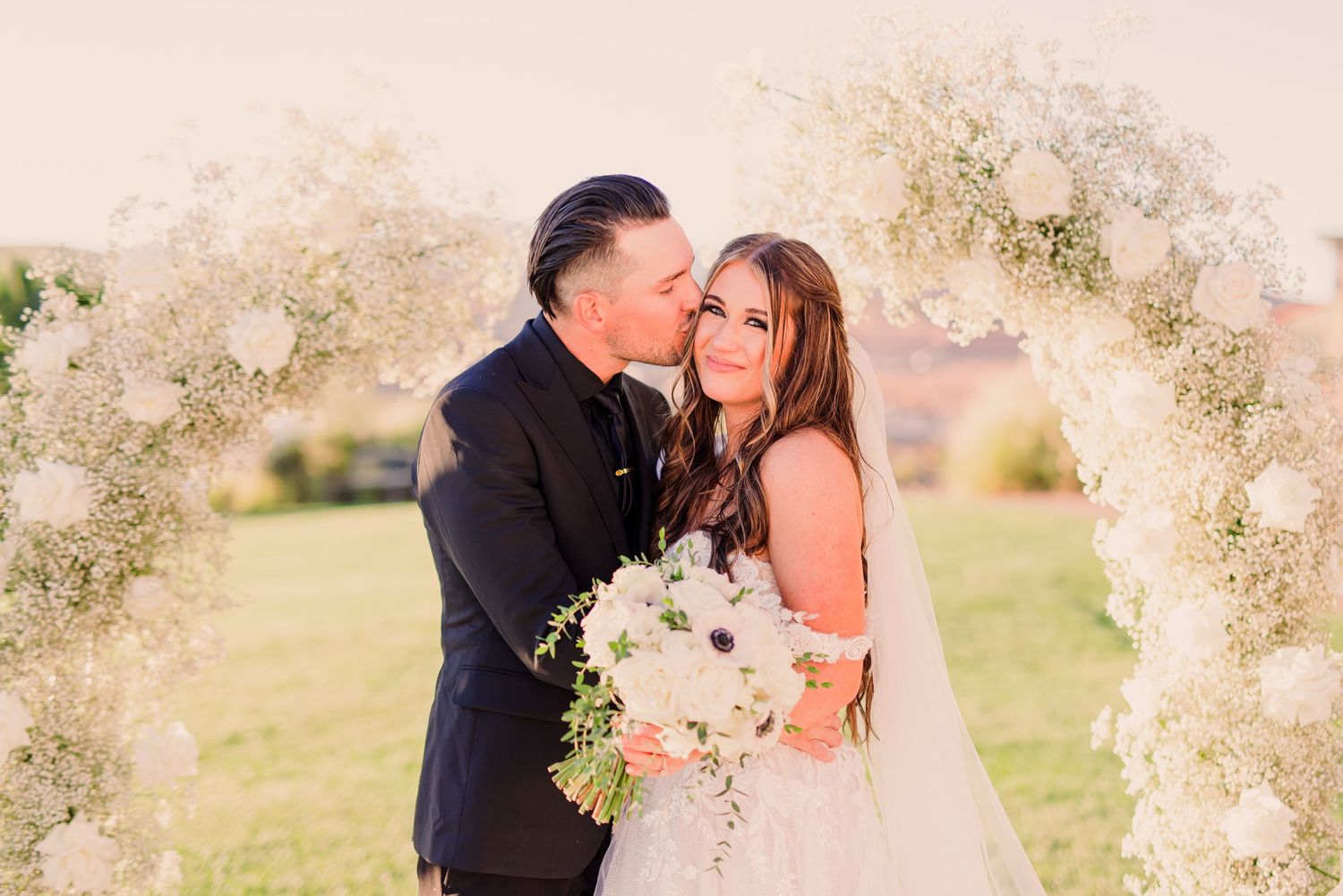 A couple in formal wedding attire walks together through a lush garden path surrounded by trees.