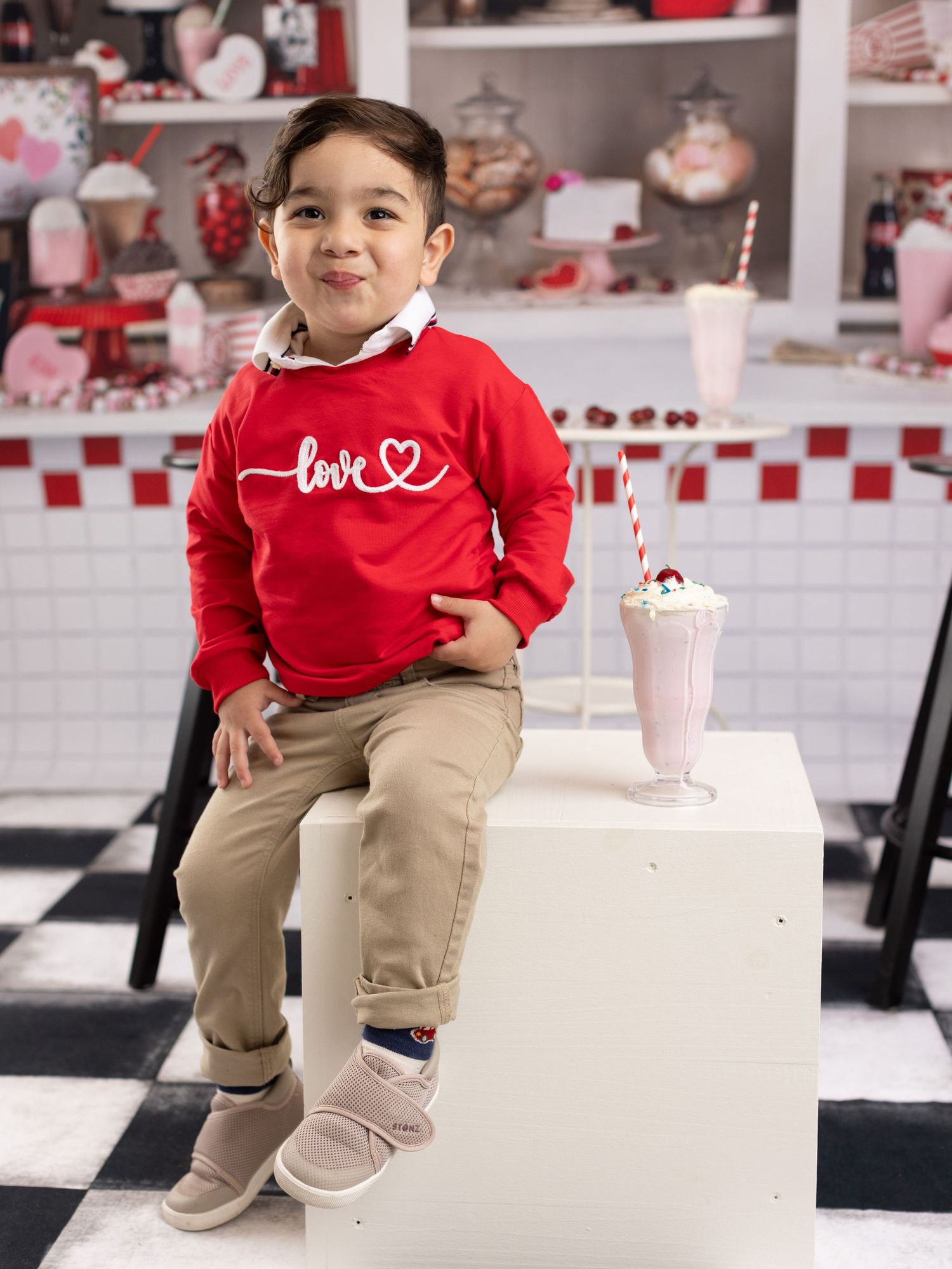 A small child in a red sweater and khaki pants sits on white steps in a Valentine's Day themed setting.