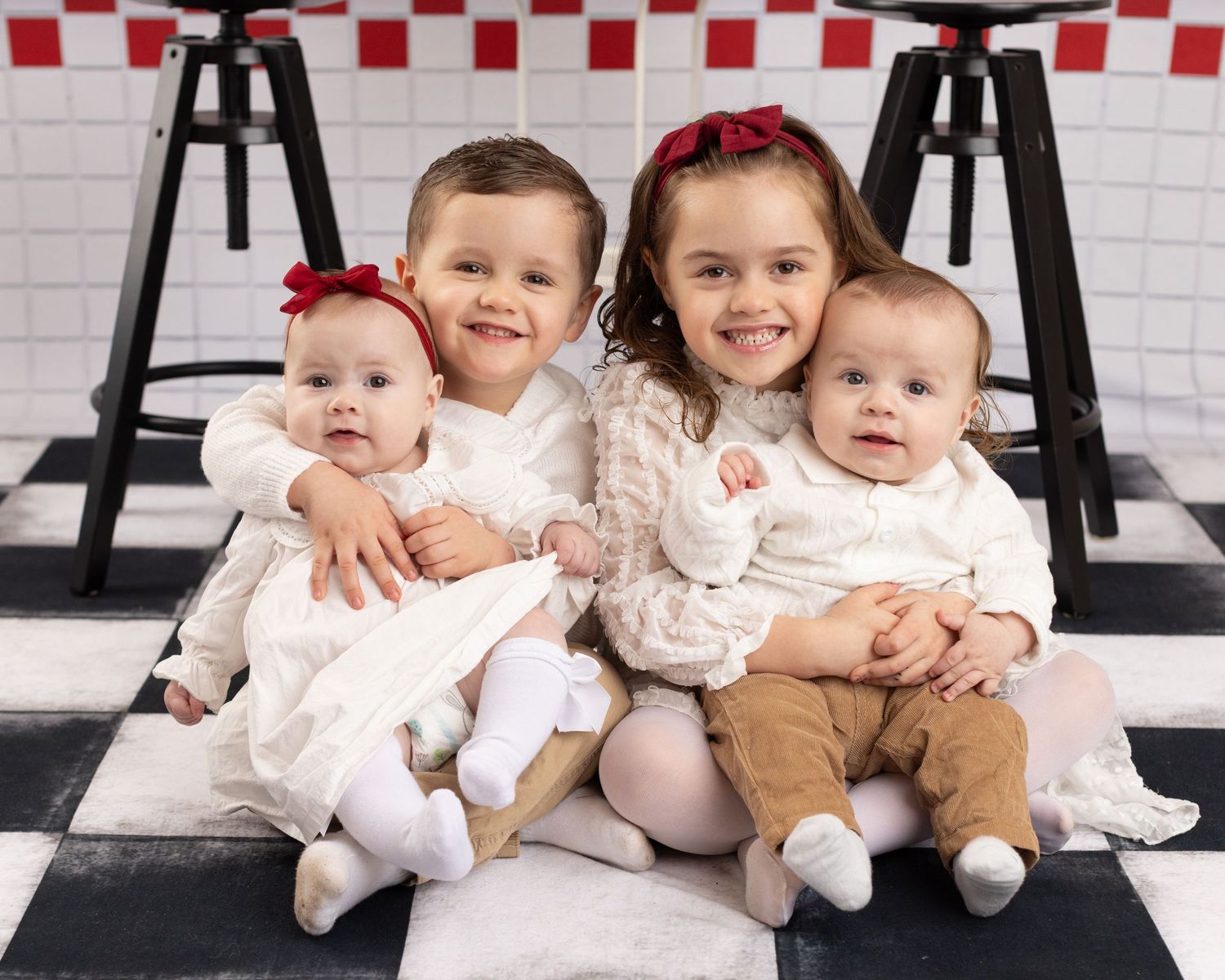 Babies and toddlers in light colored outfits sit together on a white bench against a checkered wall.